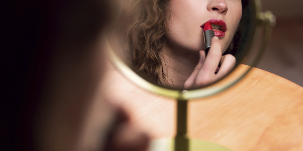 Young Woman Looking At Mirror Applying Lipstick